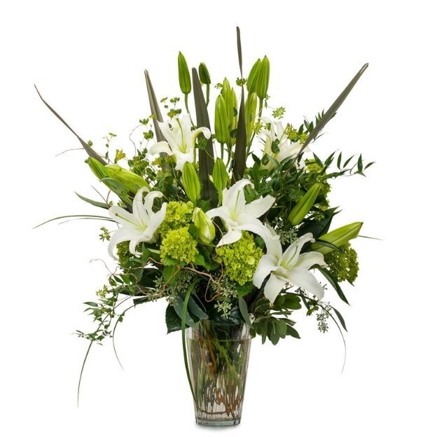 White lily bouquet with green accent flowers, unopened buds, and long leaves arranged in a clear glass vase.